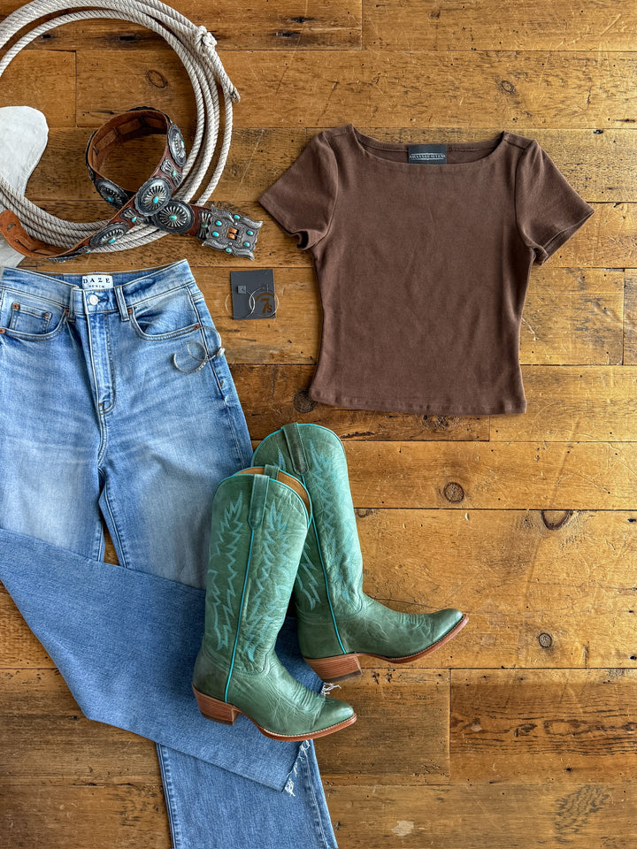 A flat lay of The Almera cocoa short sleeve top by Savannah Sevens western life{&}style, paired with buttery soft light blue jeans, green cowboy boots, a wide tooled leather belt with turquoise details, a ring, and earrings on a wooden floor.