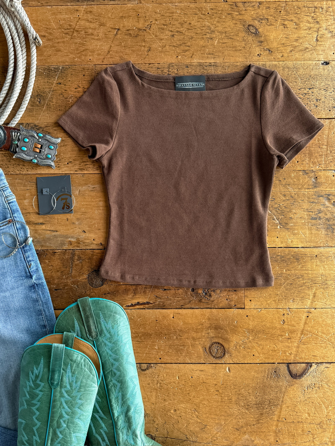 The Almera cocoa short sleeve top by Savannah Sevens western life{&}style, crafted from buttery soft tri-blend fabric, is displayed on wood beside turquoise cowboy boots, blue jeans, a leather belt with turquoise buckle, gray earring card, and coiled rope.