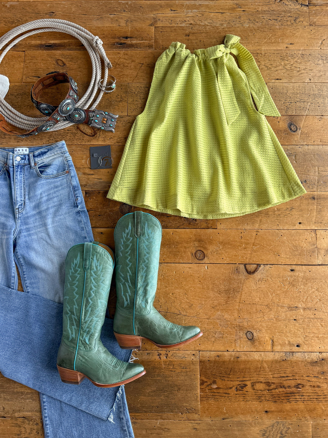 A flat lay featuring The Kalewa by Savannah Sevens western life{&}style: a green skirt with tie belt, light blue jeans, green cowboy boots, chartreuse lime top, brown belt with turquoise conchos, and silver necklace on a wooden floor.