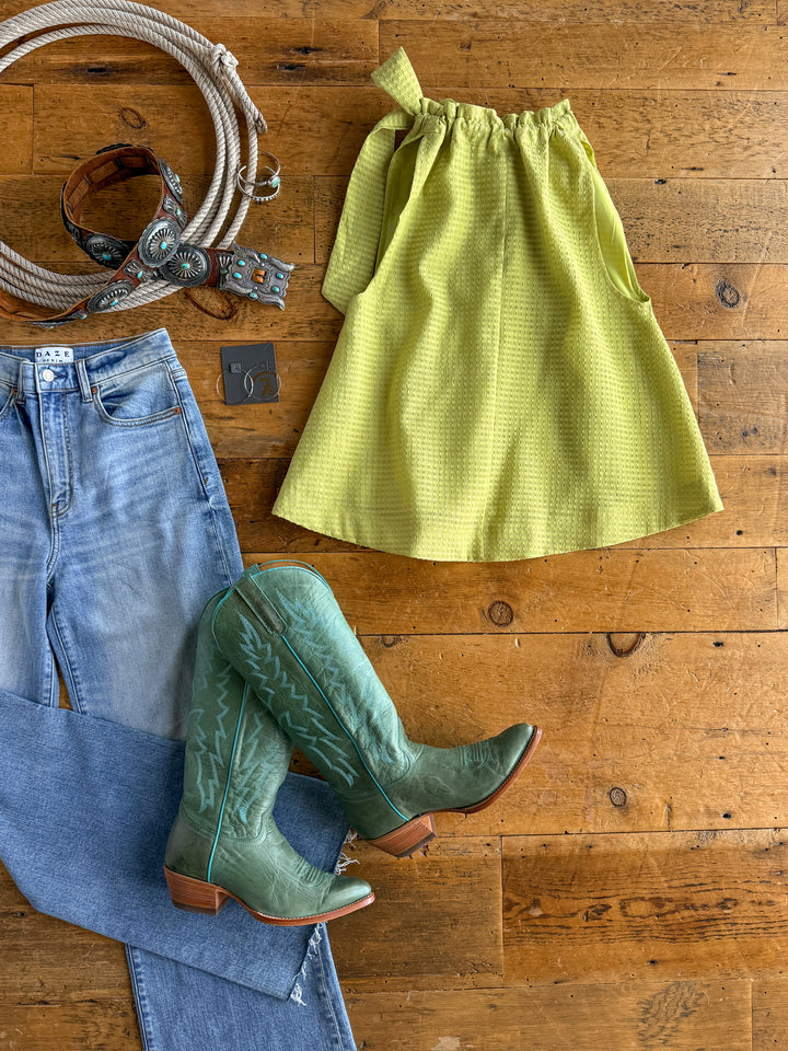 Flat lay of western items on wood floor: The Kalewa yellow tri-blend skirt by Savannah Sevens western life{&}style, blue jeans, green cowboy boots, brown leather belt with turquoise accents, coiled rope, and turquoise earrings.