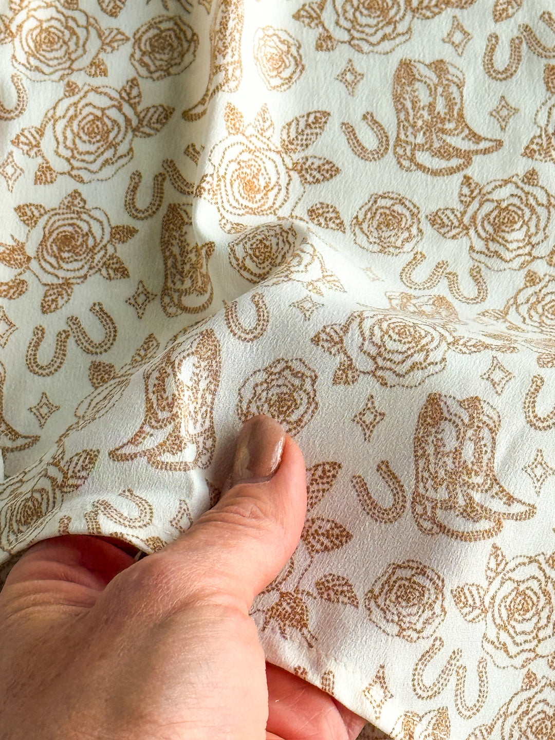 A hand holds The Nekoma {S-XXL} by Savannah Sevens western life{&}style—a white fabric with brown roses, horseshoes, and cowboy hats in a repeating design, featuring a subtle texture and soft sheen. Ideal for Western graphic tops or long sleeve button-ups.