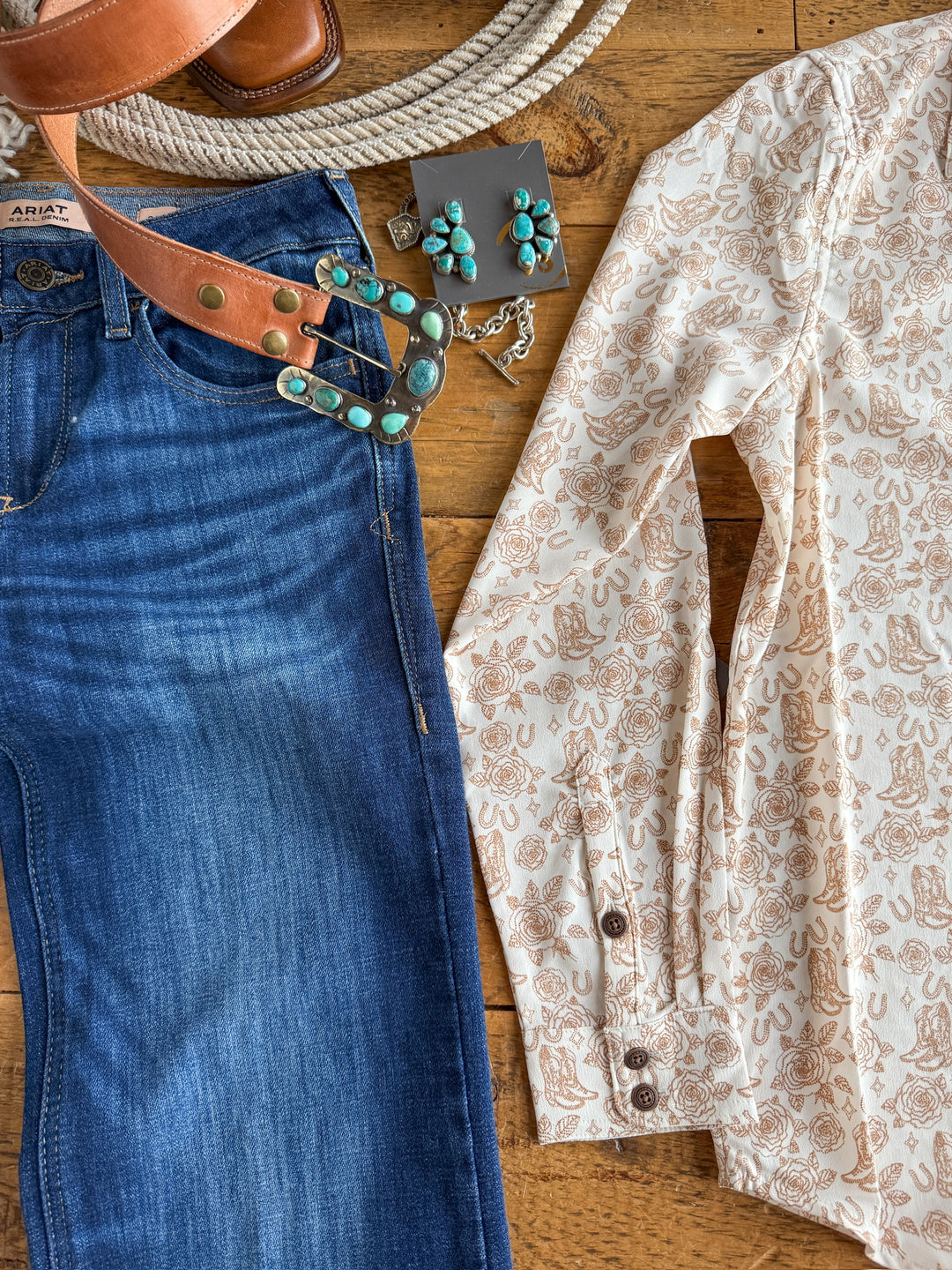Flat lay featuring The Nekoma {S-XXL} by Savannah Sevens western life{&}style: blue jeans, Western graphic top, tan leather belt with turquoise buckle, turquoise jewelry, and brown purse on a wooden surface.