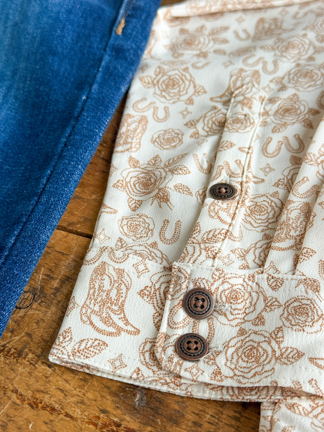 A close-up of The Nekoma top by Savannah Sevens western life{&}style, featuring a cream color with brown cowboy graphics and buttons, displayed beside blue denim on a wooden surface.