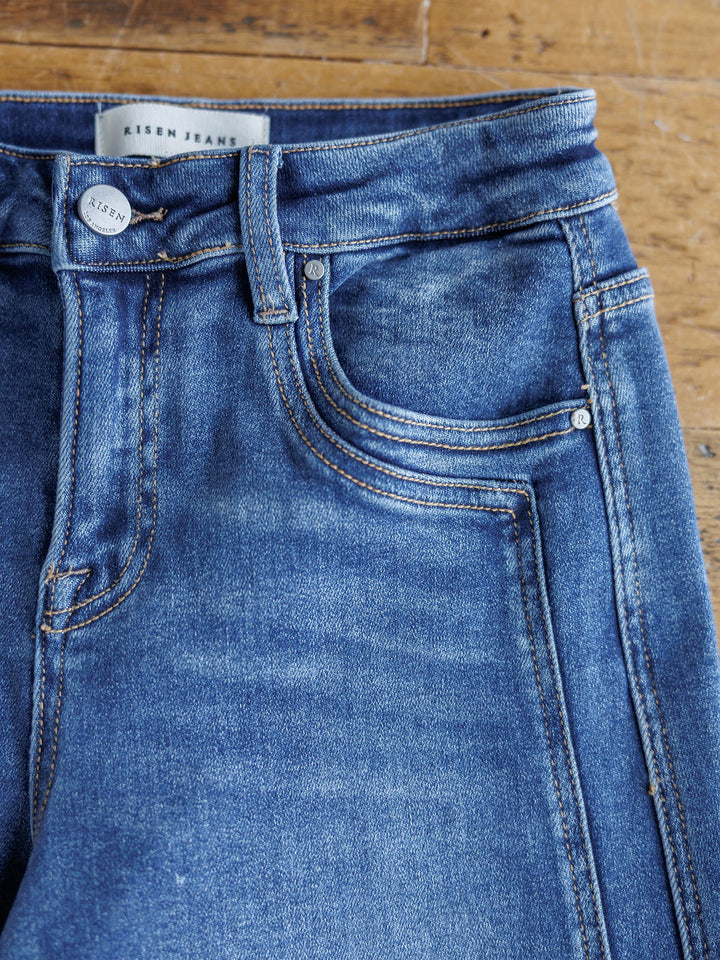 Close-up of The Maren High Rise Wide Leg Jean by Savannah Sevens western life{&}style, featuring visible stitching, front and coin pockets, belt loops, and a metal button. Jeans are laid flat on a wooden surface.