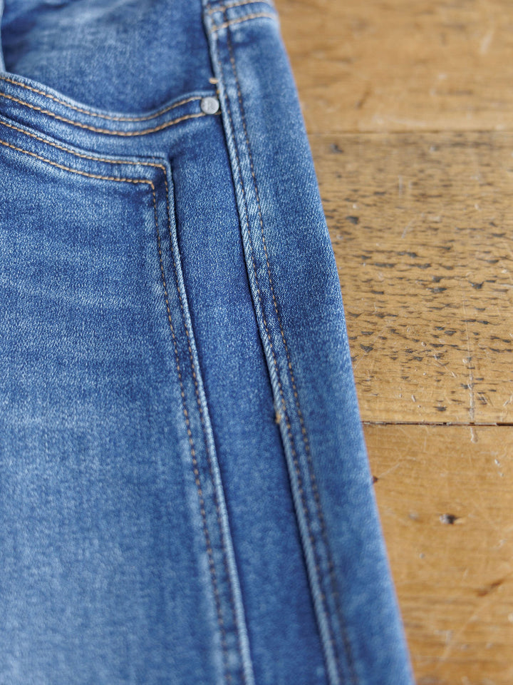A close-up of The Maren High Rise Wide Leg Jean by Savannah Sevens western life{&}style shows visible stitching on the pocket and upper leg area as the denim lies on a wooden surface.