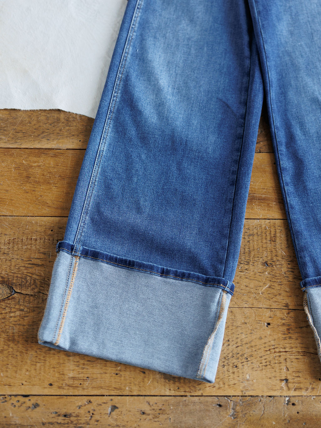 A close-up of The Maren High Rise Wide Leg Jean by Savannah Sevens western life{&}style, featuring rolled cuffs that reveal the lighter inner fabric, displayed flat on a wooden surface.
