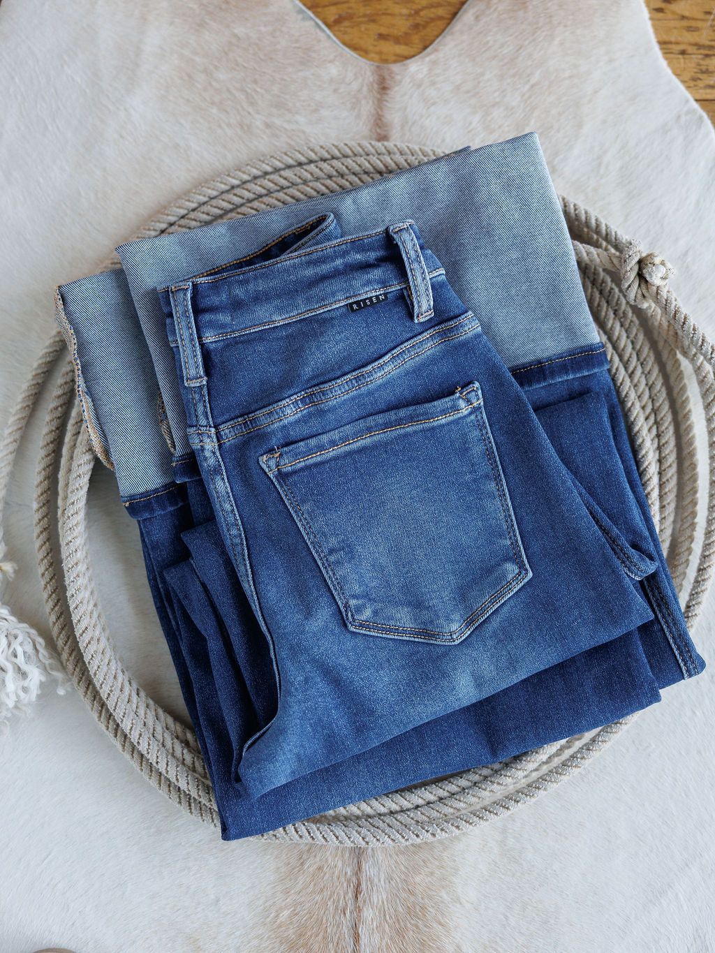 Two pairs of blue jeans, including The Maren High Rise Wide Leg Jean by Savannah Sevens western life{&}style, are folded and stacked on a coiled rope atop a light-colored animal hide rug.