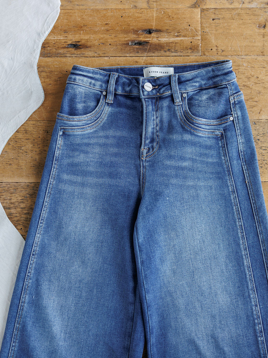 The Maren High Rise Wide Leg Jean by Savannah Sevens western life{&}style, featuring front pockets and belt loops, is laid flat on a wooden surface next to a white fabric.