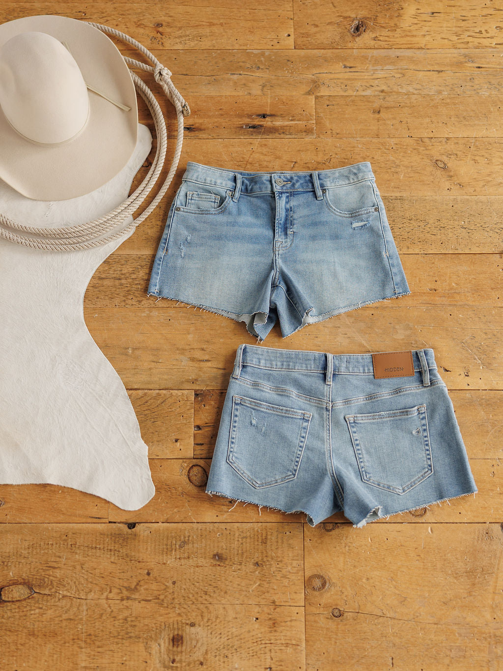 The Taylin Shorts by Savannah Sevens western life{&}style are light blue premier rise denim with a raw cut hem, shown front and back on wood flooring beside a beige cowboy hat, coiled rope, and pale hide.