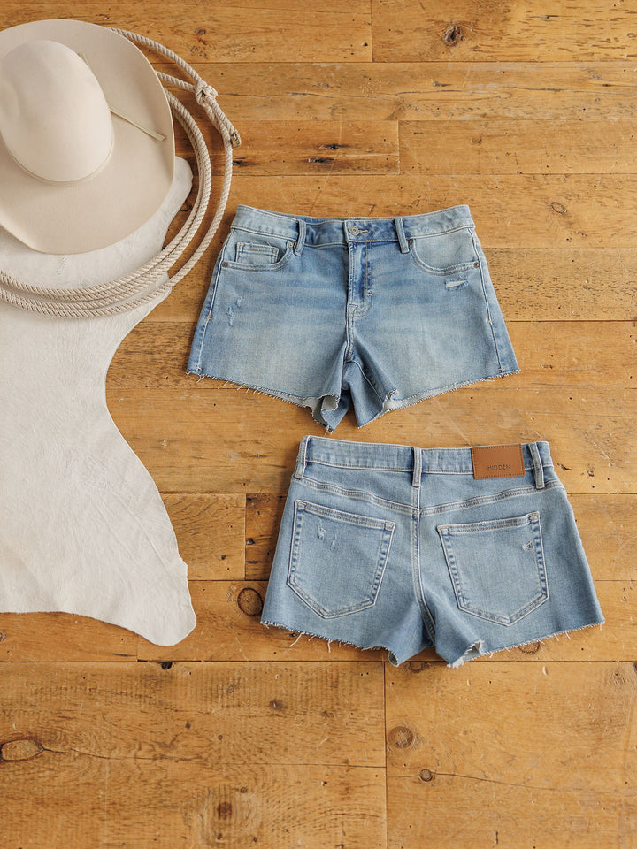 The Taylin Shorts by Savannah Sevens western life{&}style are light blue premier rise denim with a raw cut hem, shown front and back on wood flooring beside a beige cowboy hat, coiled rope, and pale hide.