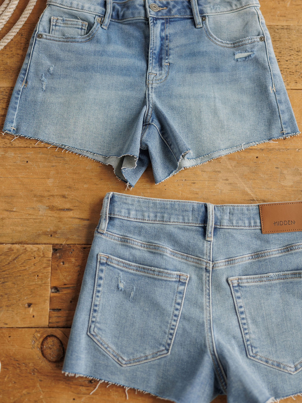 Two pairs of The Taylin Shorts by Savannah Sevens western life{&}style, in a medium light wash with raw cut hems, are displayed flat on wood; one shows the front and the other the back with pockets and a brown leather patch.