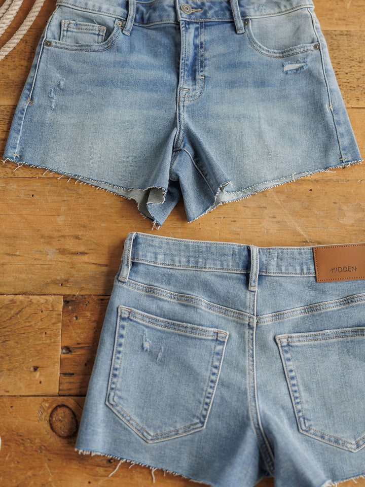Two pairs of The Taylin Shorts by Savannah Sevens western life{&}style, in a medium light wash with raw cut hems, are displayed flat on wood; one shows the front and the other the back with pockets and a brown leather patch.