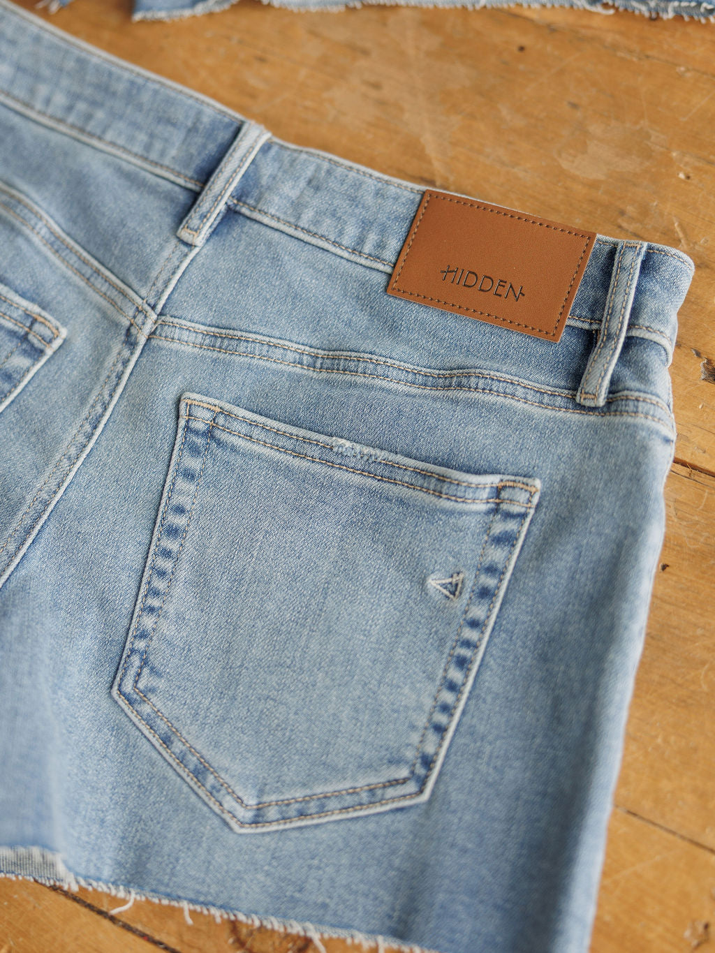 The Taylin Shorts by Savannah Sevens western life{&}style: light blue rise denim shorts with raw cut hems, back pockets, and a brown HIDDEN leather patch on the waistband, shown on a wooden surface.