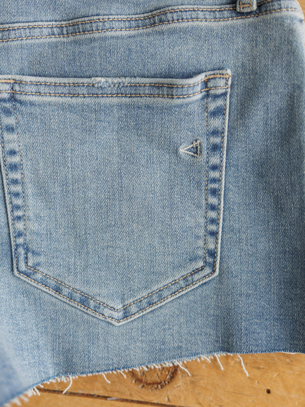 Close-up of The Taylin Shorts by Savannah Sevens western life{&}style in medium light wash denim, with visible stitching, a small triangular back pocket detail, and raw cut hem. Shown on a wooden surface.