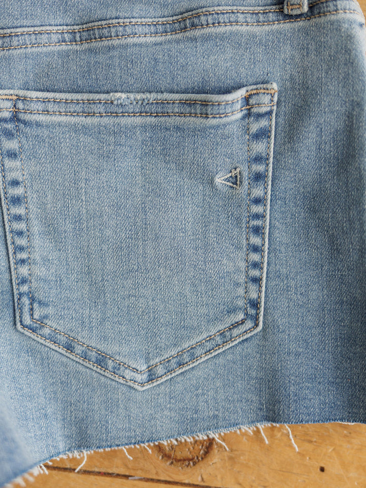 Close-up of The Taylin Shorts by Savannah Sevens western life{&}style in medium light wash denim, with visible stitching, a small triangular back pocket detail, and raw cut hem. Shown on a wooden surface.
