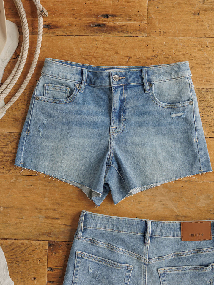 The Taylin Shorts by Savannah Sevens western life{&}style are medium light wash denim shorts with a raw cut hem and subtle distressing, shown flat on wood next to another pair of shorts and a coiled rope.