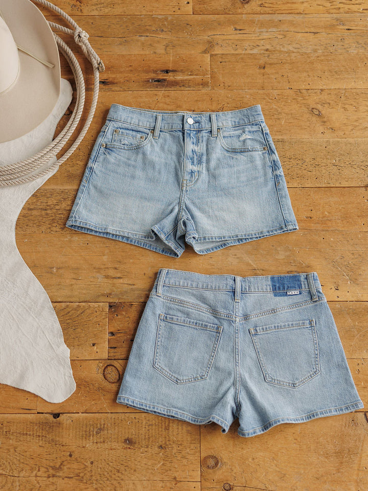 Two pairs of The Sutton Shorts by Savannah Sevens western life{&}style in medium light wash, front and back, laid on wood beside a beige cowboy hat, rope, and light fabric. Made from organic cotton for comfort and style.