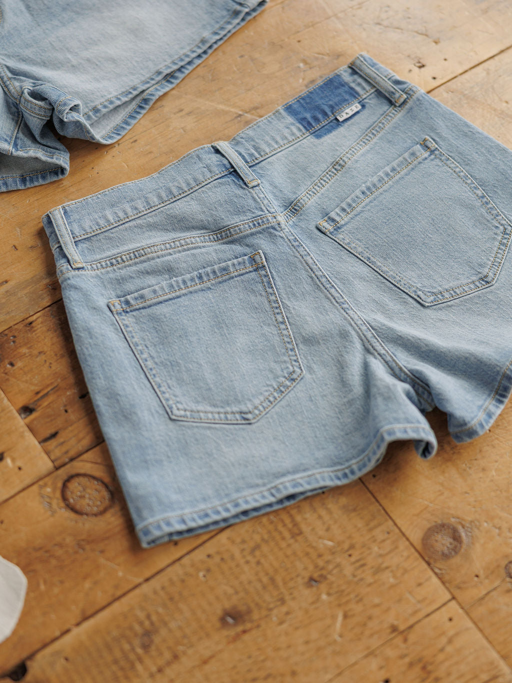 The Sutton Shorts by Savannah Sevens western life{&}style in a medium light wash are laid flat, displaying the back pockets and waistband. Another pair of denim shorts is partially visible in the corner.
