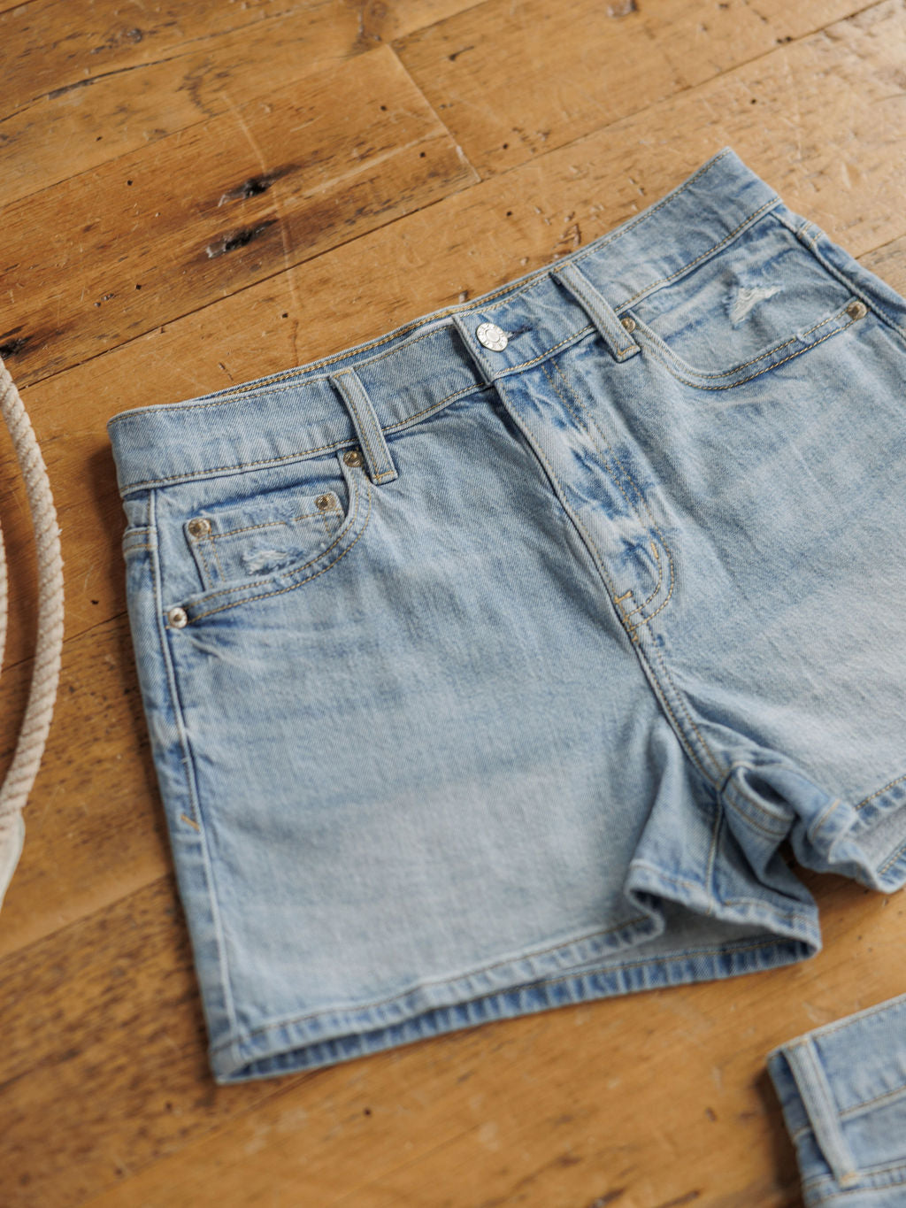 The Sutton Shorts by Savannah Sevens western life{&}style are light blue high rise denim shorts with a medium light wash, front pockets, and button fly, displayed flat on a wooden floor with a rope partially visible on the left.