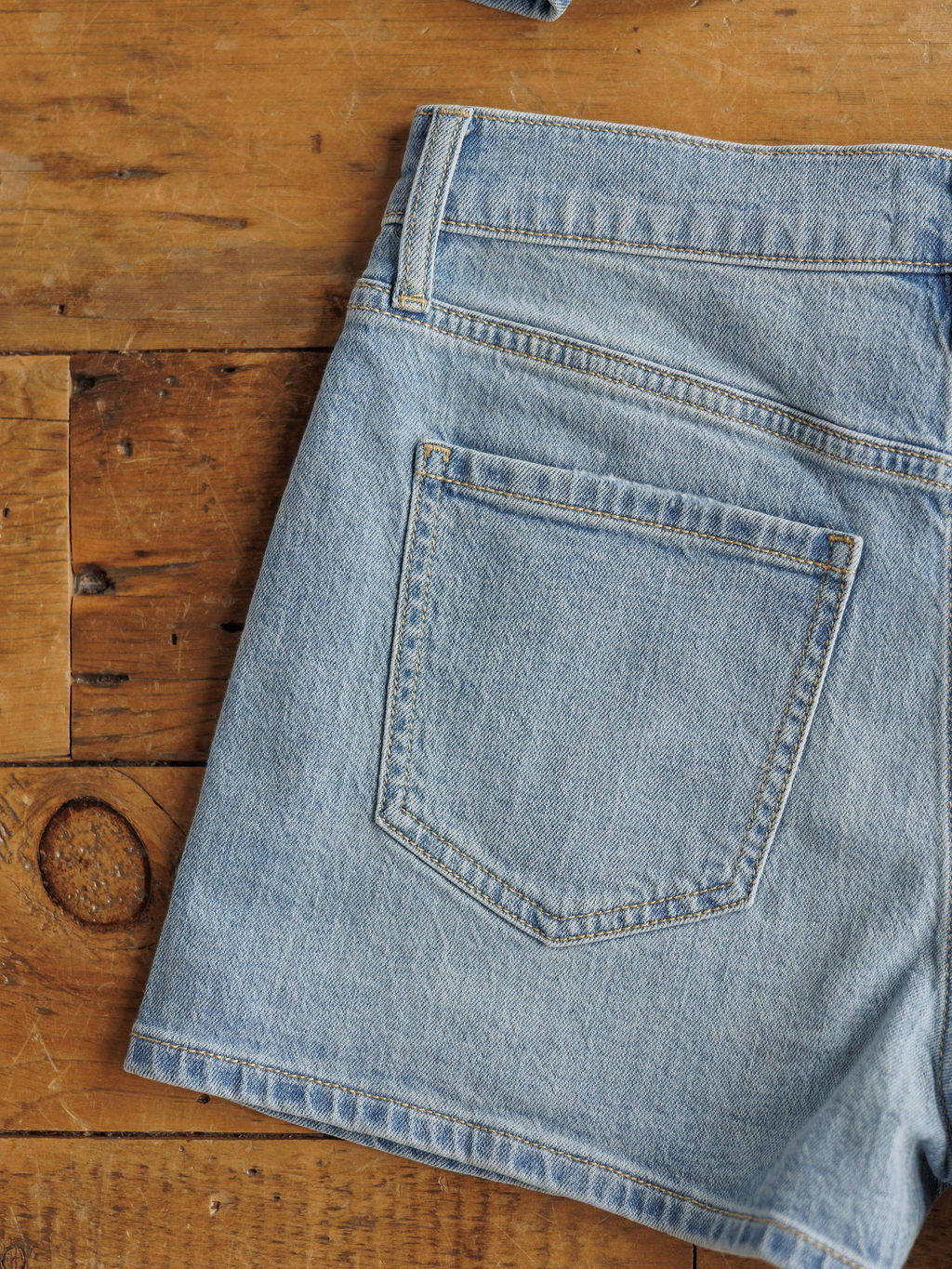 A close-up of The Sutton Shorts by Savannah Sevens western life{&}style, featuring high-rise, medium light wash organic cotton denim with a back pocket and waistband detail on a rustic wooden surface.