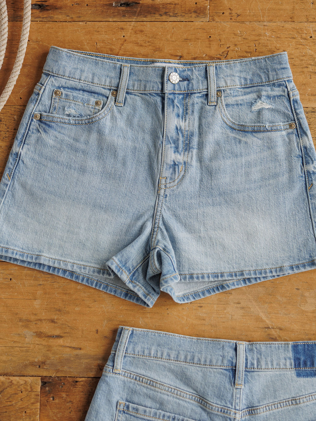 The Sutton Shorts by Savannah Sevens western life{&}style are medium light wash, high-rise denim shorts with front pockets and a button closure, laid flat on wood. Another similar pair is partially visible at the image bottom.