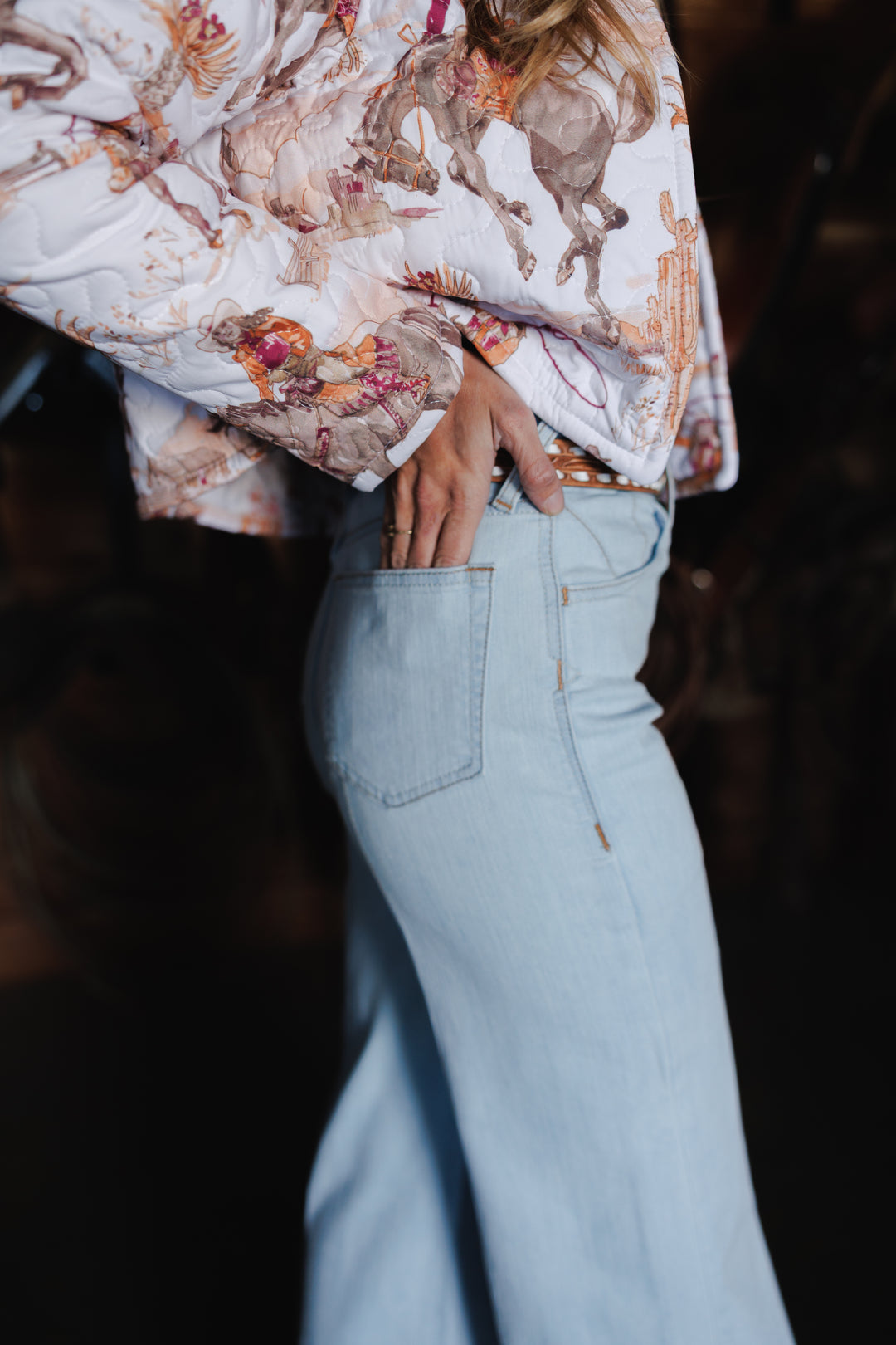 A person wearing light blue jeans and a long-sleeved shirt with a horse print rests their hand in their back pocket, shown from the waist down and side view, embodying casual Western style.