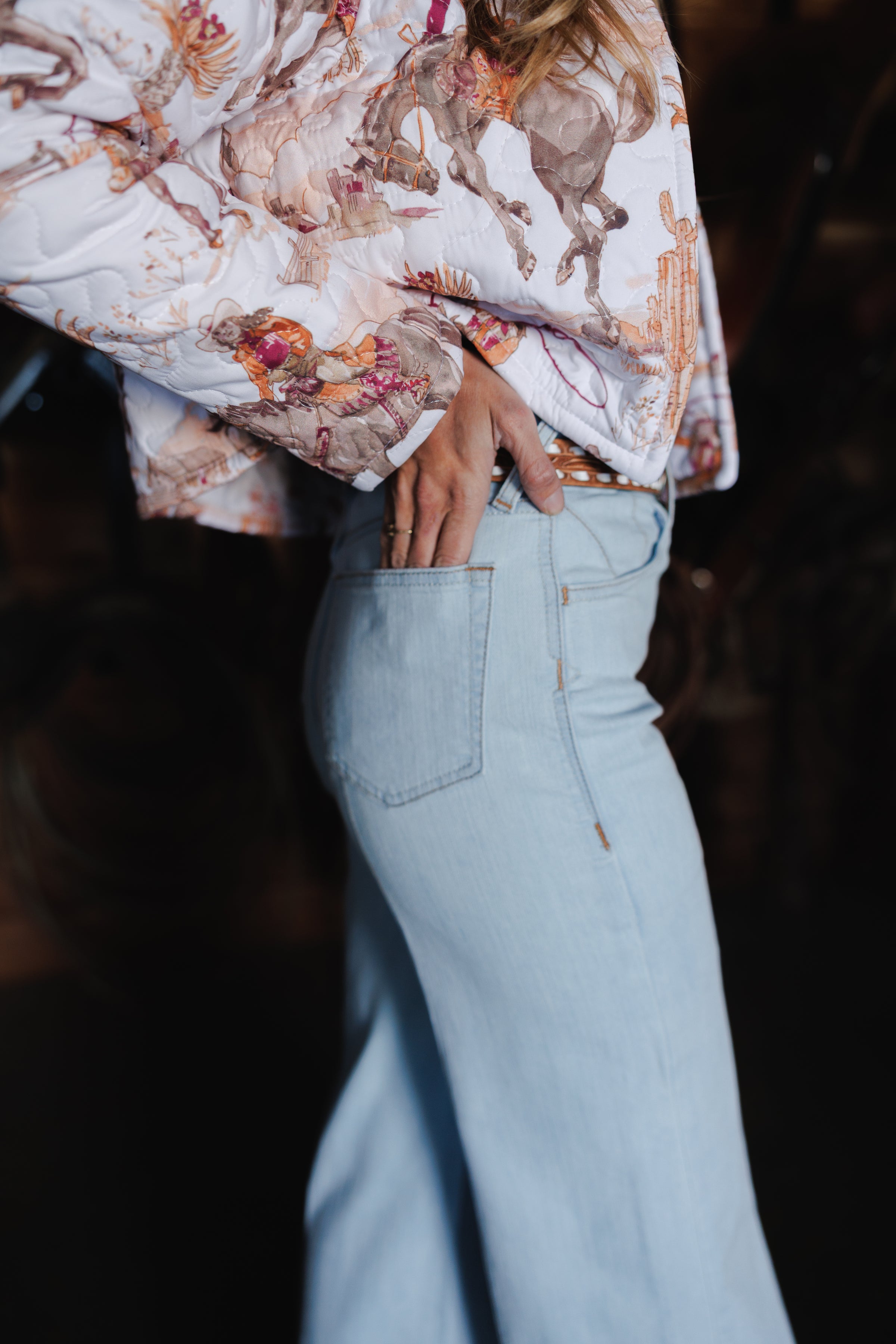 A person wearing light blue jeans and a long-sleeved shirt with a horse print rests their hand in their back pocket, shown from the waist down and side view, embodying casual Western style.