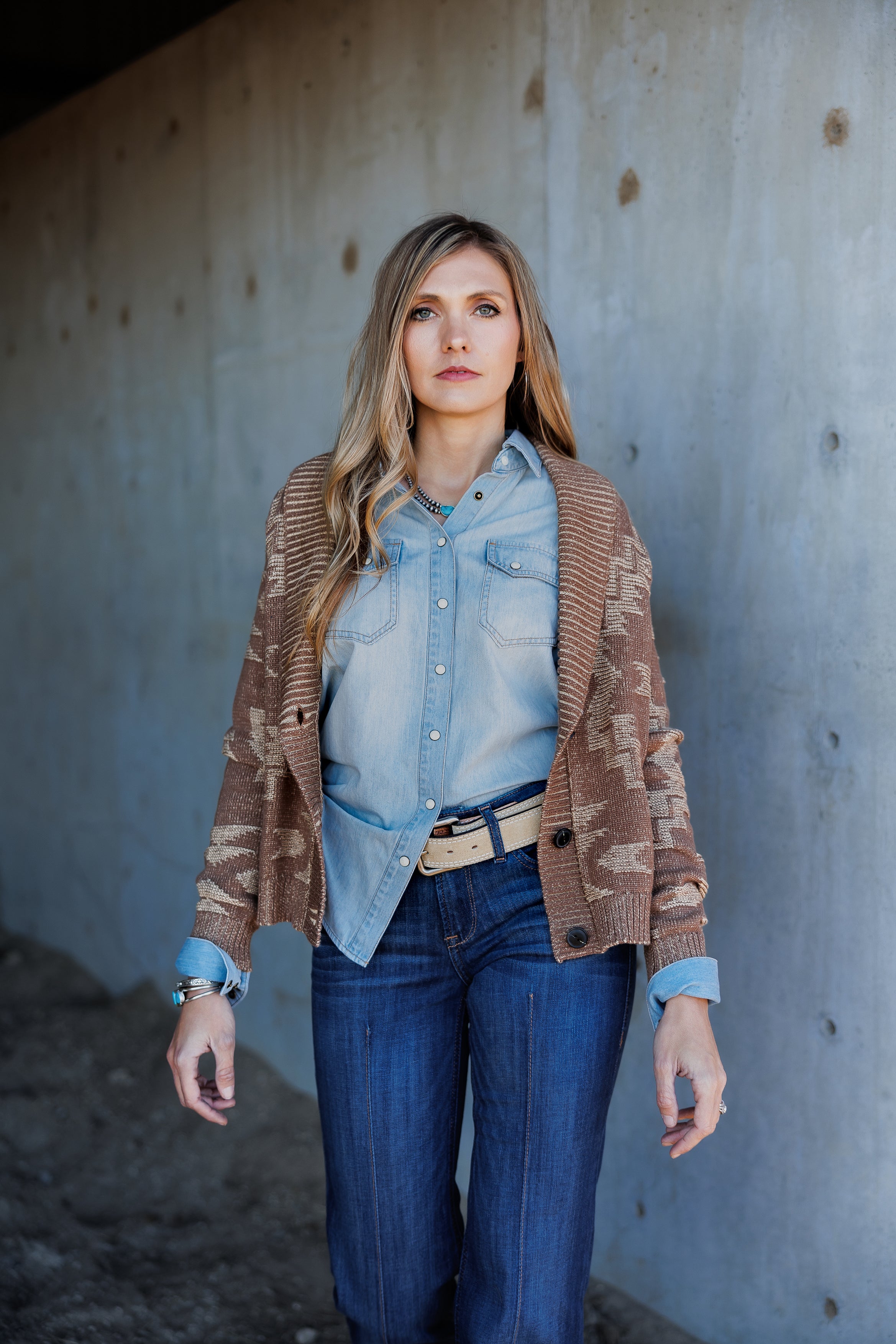 A woman with long blonde hair stands in front of a concrete wall, wearing a light denim shirt, blue jeans, a beige belt, and a brown patterned cardigan. She looks ahead with a neutral expression, her relaxed style enhanced by the classic denim ensemble.