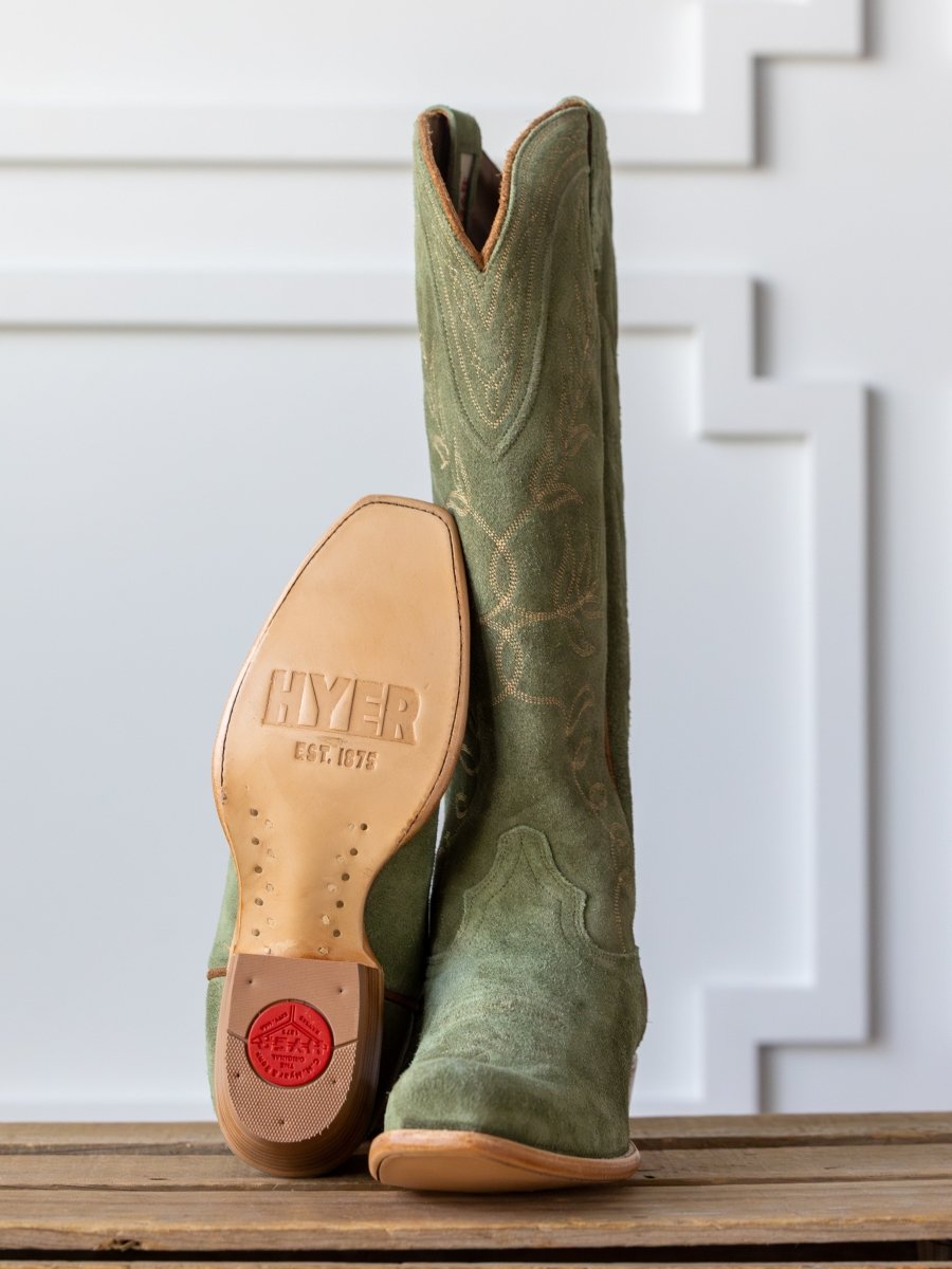 A pair of green suede The Hyer Abbyville Boot by Savannah Sevens western life{&}style—one upright, the other showing its branded sole and red heel cap—displayed on a wooden surface before a white decorative wall.