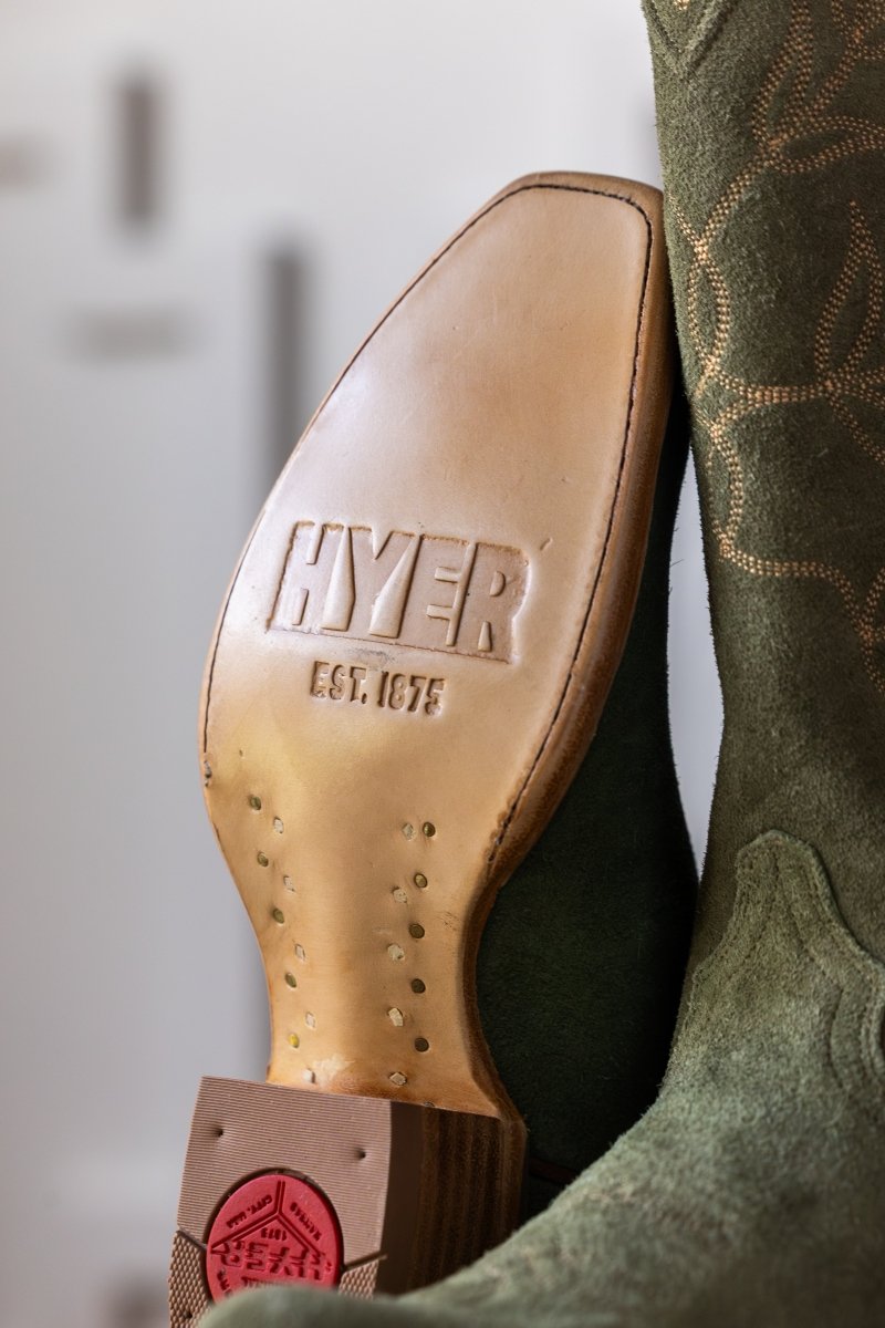 Close-up of the sole of The Hyer Abbyville Boot by Savannah Sevens western life{&}style. "HYER EST. 1875" is embossed on the leather sole, with part of the green suede upper and decorative stitching visible.