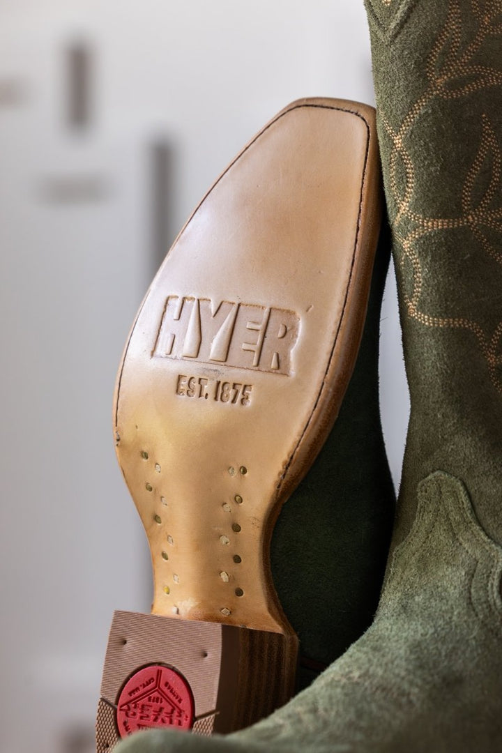 Close-up of the sole of The Hyer Abbyville Boot by Savannah Sevens western life{&}style. "HYER EST. 1875" is embossed on the leather sole, with part of the green suede upper and decorative stitching visible.
