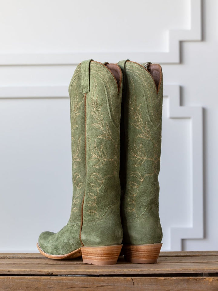 The  Hyer Abbyville Boot by Savannah Sevens western life{&}style—a pair of tall, green roughout leather boots with tan embroidery and wooden heels—is showcased on a wooden surface against a white paneled background.