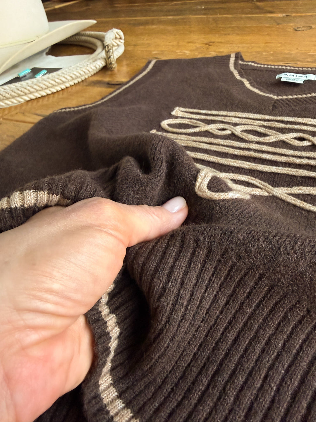 A hand pinches the soft tri-blend knit fabric of Savannah Sevens western life{&}style’s Abilene Boot Stitch Sweater Vest, featuring chocolate knit with tan patterns, laid on a wooden surface. A cowboy hat and rope can be seen in the background.