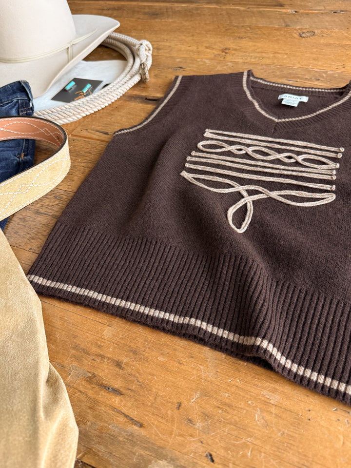 The Abilene Boot Stitch Sweater Vest by Savannah Sevens western life{&}style, a chocolate knit vest featuring beige decorative stitching, is displayed on a wooden table alongside a white cowboy hat, denim jeans, a tan belt, and a tan suede accessory in the corner.