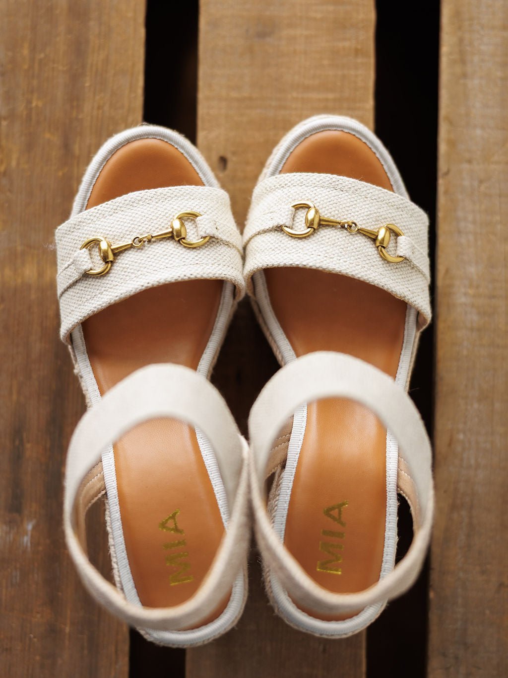 The Abilene by Savannah Sevens western life{&}style: tan wedge sandals with open toe and gold snaffle bit details on the straps, displayed on wood. The insoles feature "MIA" in gold letters.