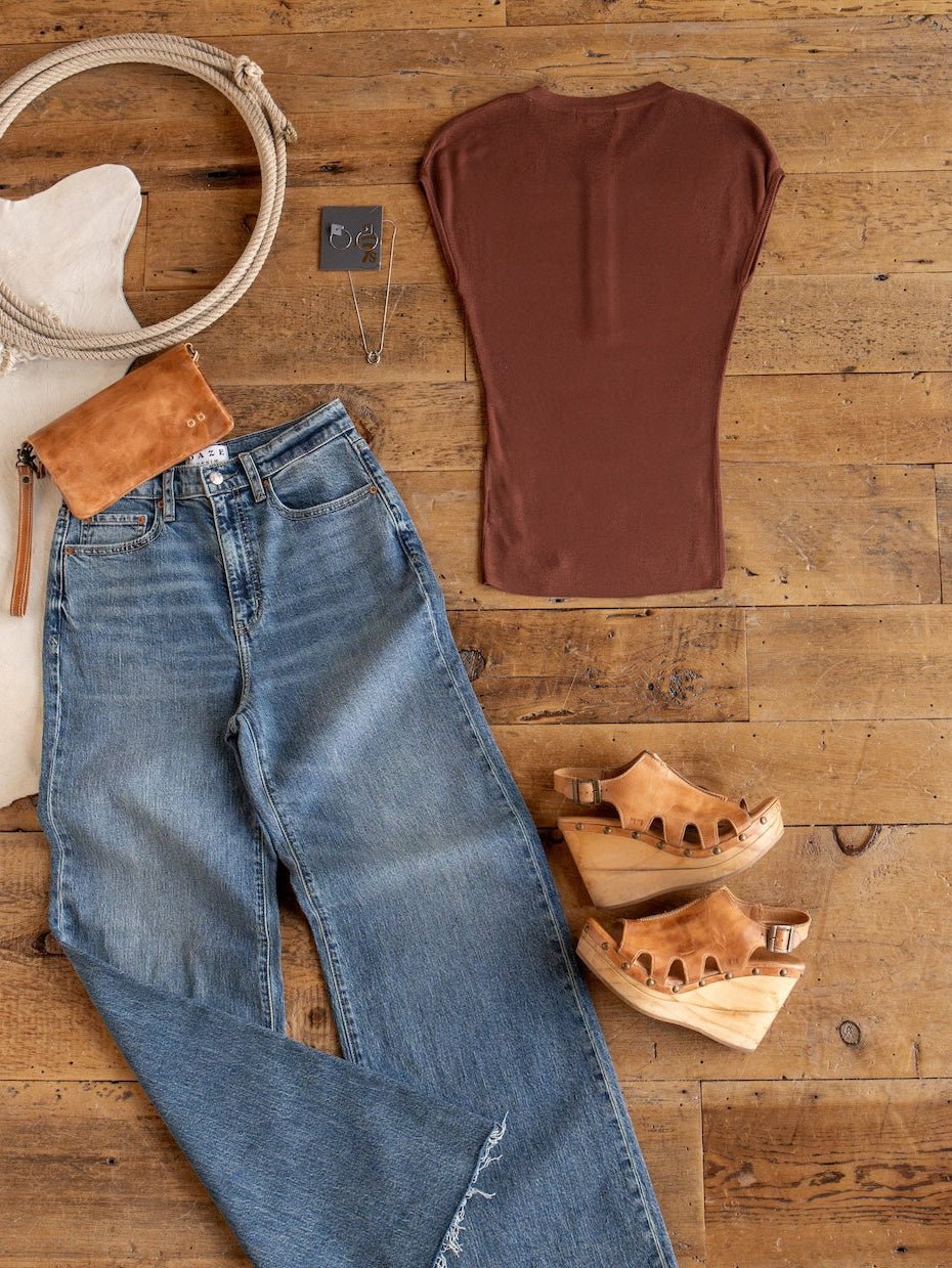 Flat lay of blue jeans, the Adair Chestnut Cap Sleeve Top by Savannah Sevens western life{&}style, tan wooden sandals, a tan clutch, a necklace, earrings, and a white hat with a rope detail on a wooden floor.