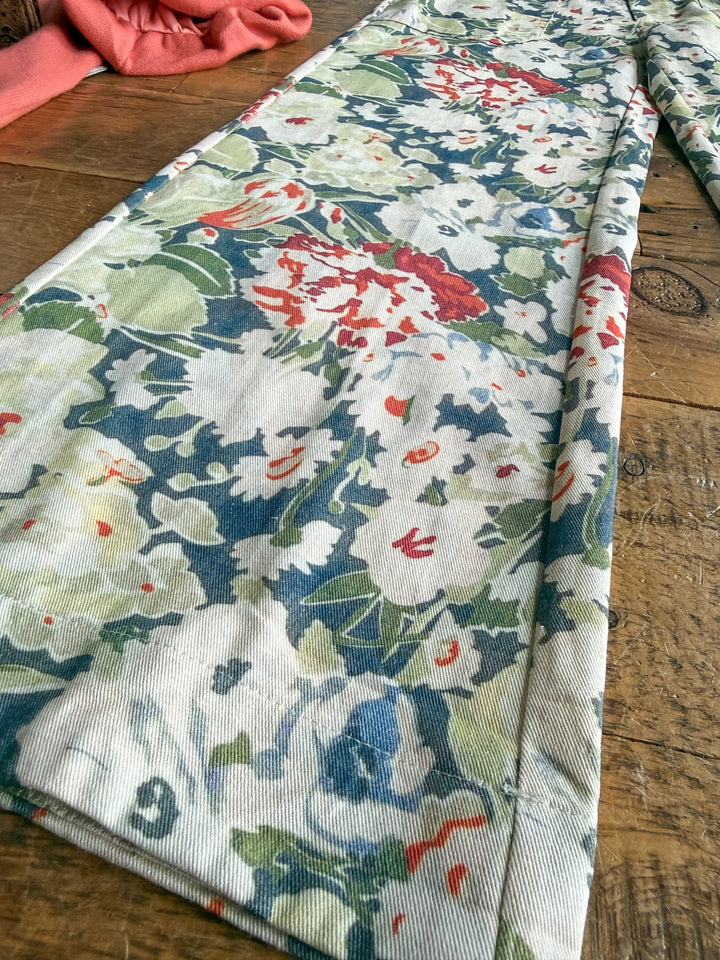 The Adelanto Floral Wide Leg Crop Pants by Savannah Sevens western life{&}style, featuring a red, green, and blue floral print, are displayed on a wooden surface with a piece of pink fabric visible in the top left corner.