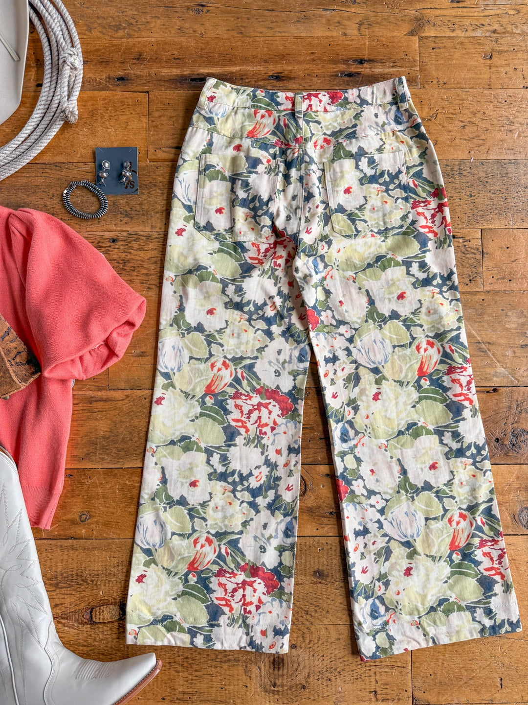 The Adelanto Floral Wide Leg Crop Pants by Savannah Sevens western life{&}style are laid out on a wooden floor, complemented by a coral sweater, white boots, a coiled rope, a pair of earrings, and a bracelet arranged nearby as part of a styled outfit display.