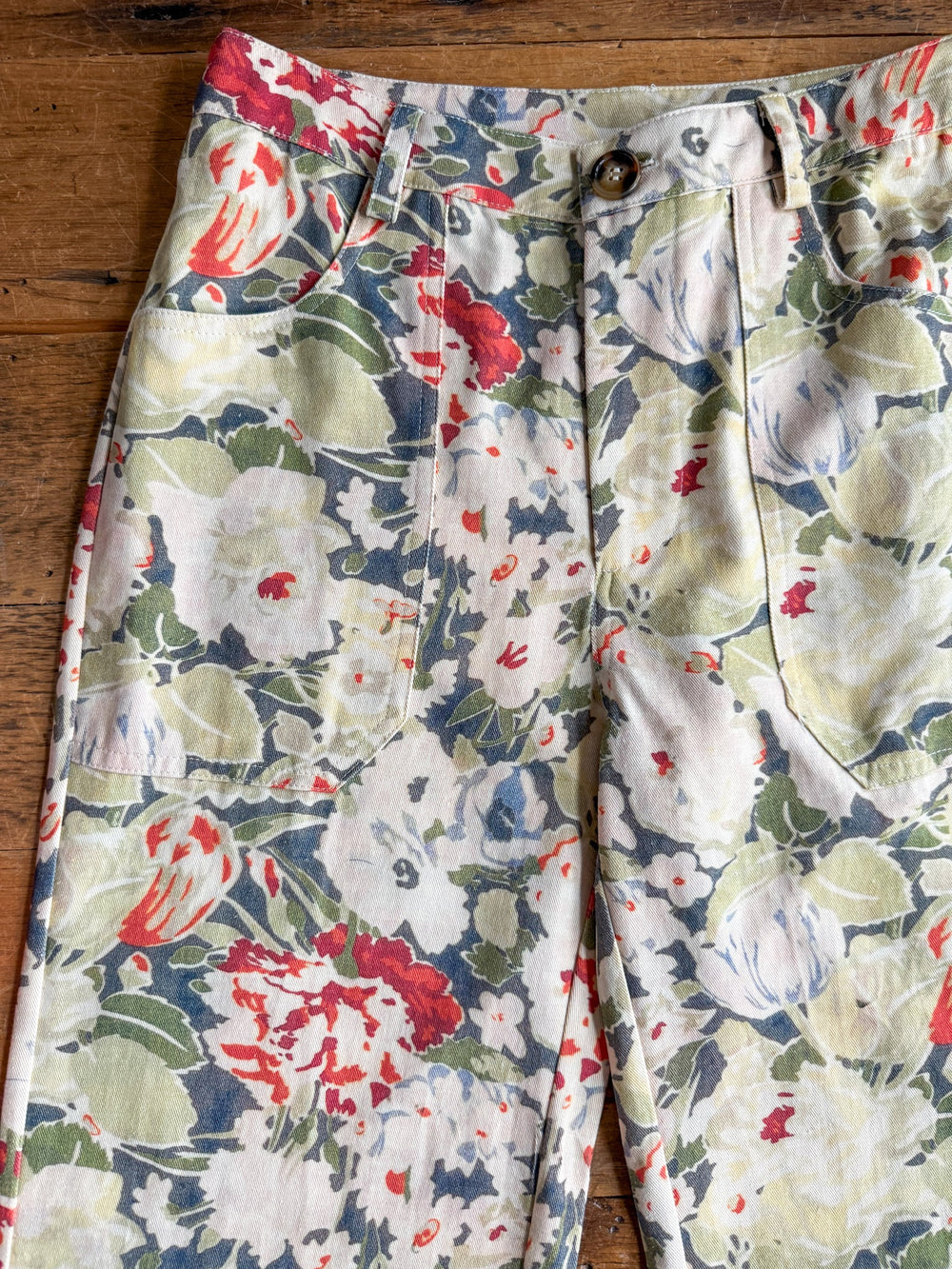 The Adelanto Floral Wide Leg Crop Pants by Savannah Sevens western life{&}style are high waisted with a wide leg silhouette, featuring a vibrant floral print in shades of green, red, white, and blue. Shown lying flat on a wooden surface, the pants highlight the waistband, pockets, and button closure.