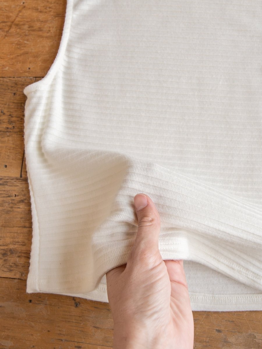 A hand stretches the ribbed fabric of the Adona Off-White Ribbed Basic Cropped Tank by Savannah Sevens western life{&}style on a wooden surface, highlighting its texture and elasticity.