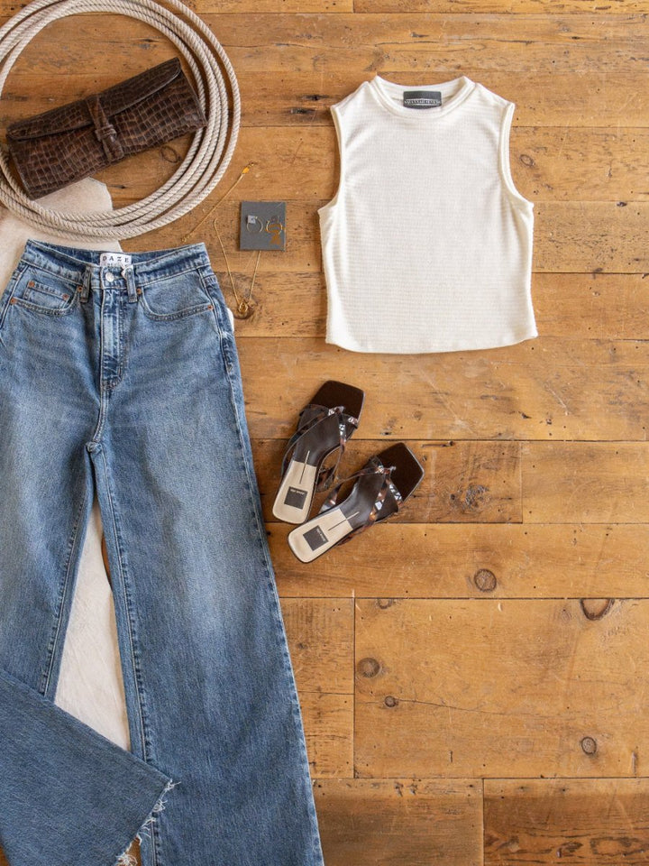 Flat lay of a casual outfit on a wooden floor featuring Savannah Sevens western life{&}style Adona Off-White Ribbed Basic Cropped Tank, blue high-waisted jeans, brown heeled sandals, brown clutch, coiled rope, earrings, and a beige cloth.