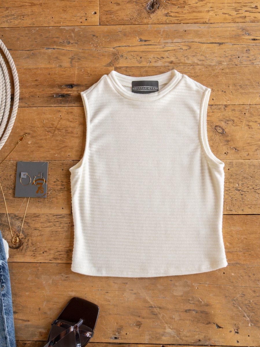 The Adona Off-White Ribbed Basic Cropped Tank by Savannah Sevens western life{&}style is laid flat on a wooden surface, shown with a necklace, shoe, partial belt, jeans, and a small card with metallic numbers nearby.