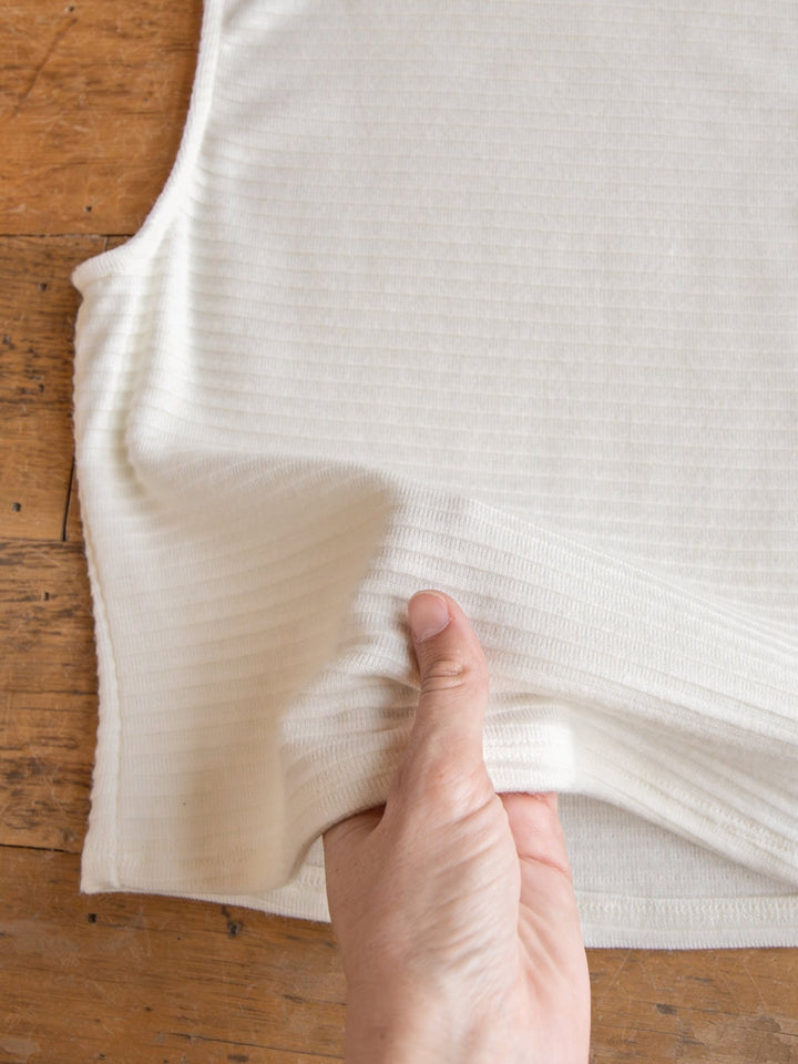 A hand stretches the ribbed fabric of the Adona Off-White Ribbed Basic Cropped Tank by Savannah Sevens western life{&}style on a wooden surface, highlighting its texture and elasticity.