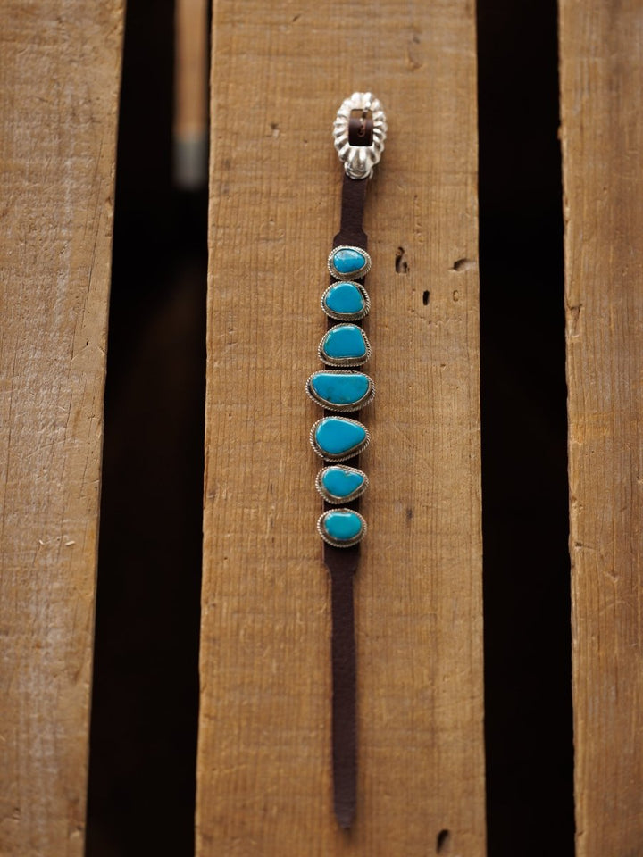 The Aguila Bracelet by Savannah Sevens western life{&}style features six genuine turquoise stones set in sterling silver on brown leather, topped with a decorative silver clasp and displayed vertically on wood.