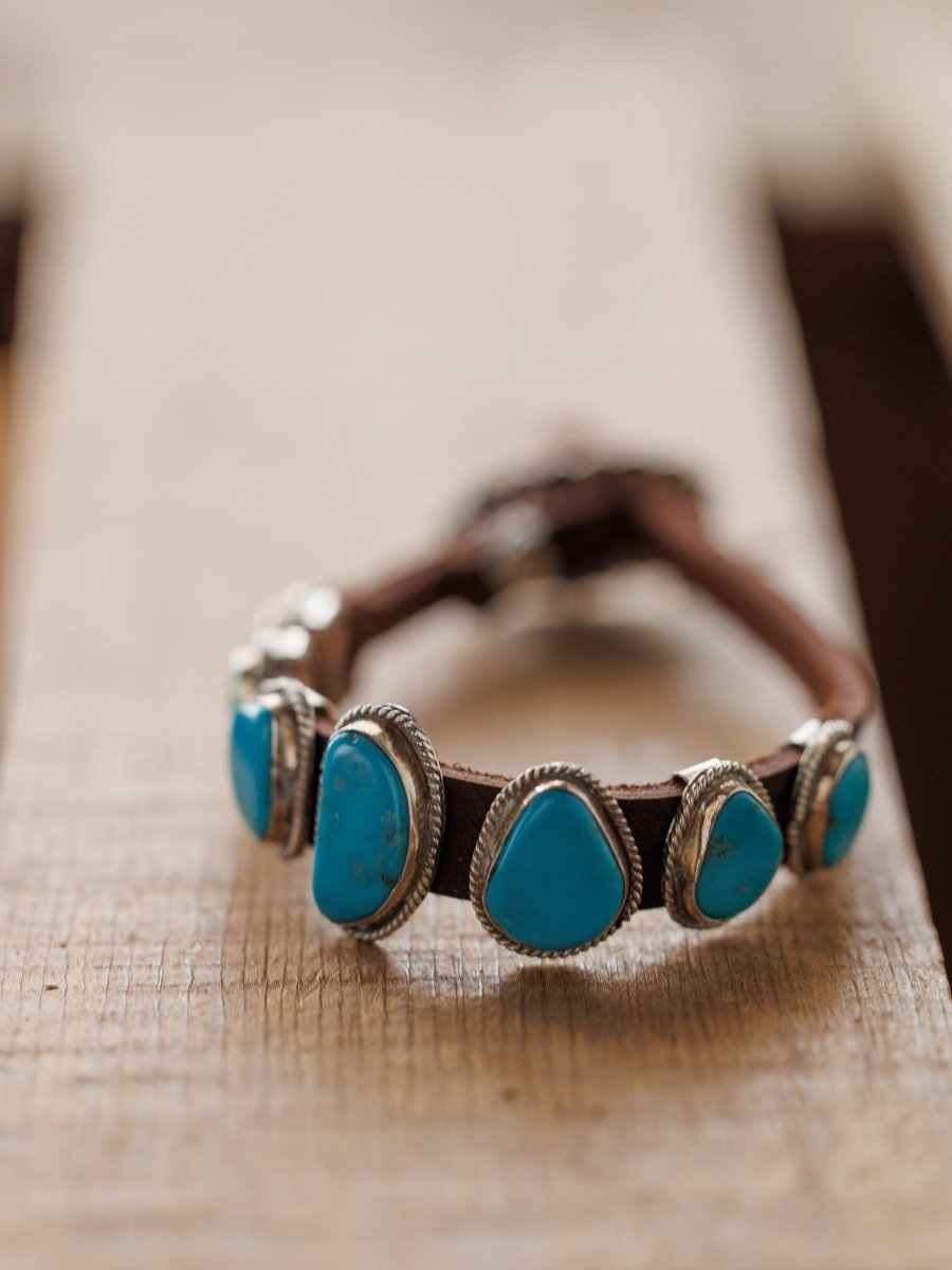 The Aguila Bracelet by Savannah Sevens western life{&}style features five genuine turquoise stones set in sterling silver on leather, displayed on a wooden surface with a blurred background.