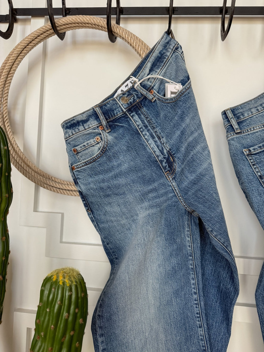 The Aiden High Rise Wide Leg Jean by Savannah Sevens western life{&}style hangs on a black metal hook, with a coiled rope and green cacti beside it, set against a white wall with decorative molding.