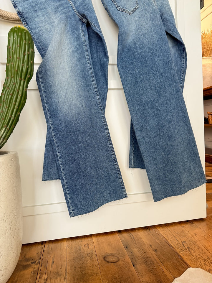 Two pairs of The Aiden High Rise Wide Leg Jeans by Savannah Sevens western life{&}style hang side by side on a white door, with an organic cotton green potted cactus nearby and warm light illuminating the wooden floor.