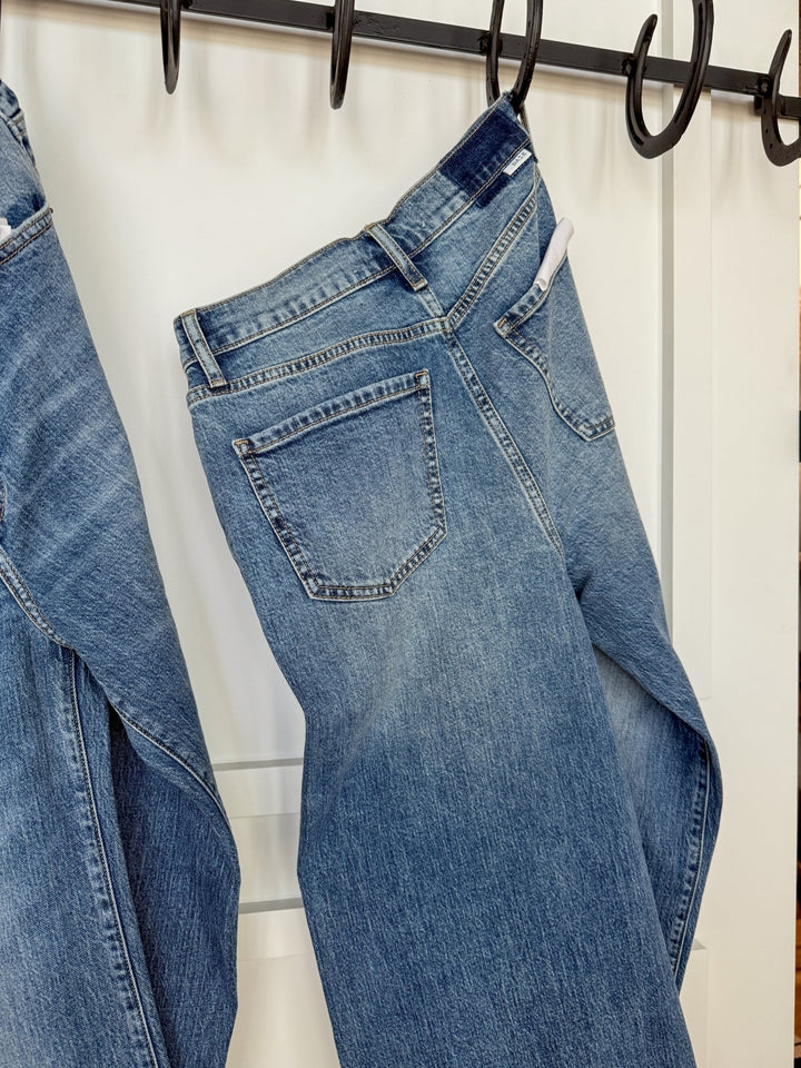 The Aiden High Rise Wide Leg Jean by Savannah Sevens western life{&}style in blue organic cotton hangs by the waistband on a black hook rack, with another pair partially visible beside it, set against a white background.