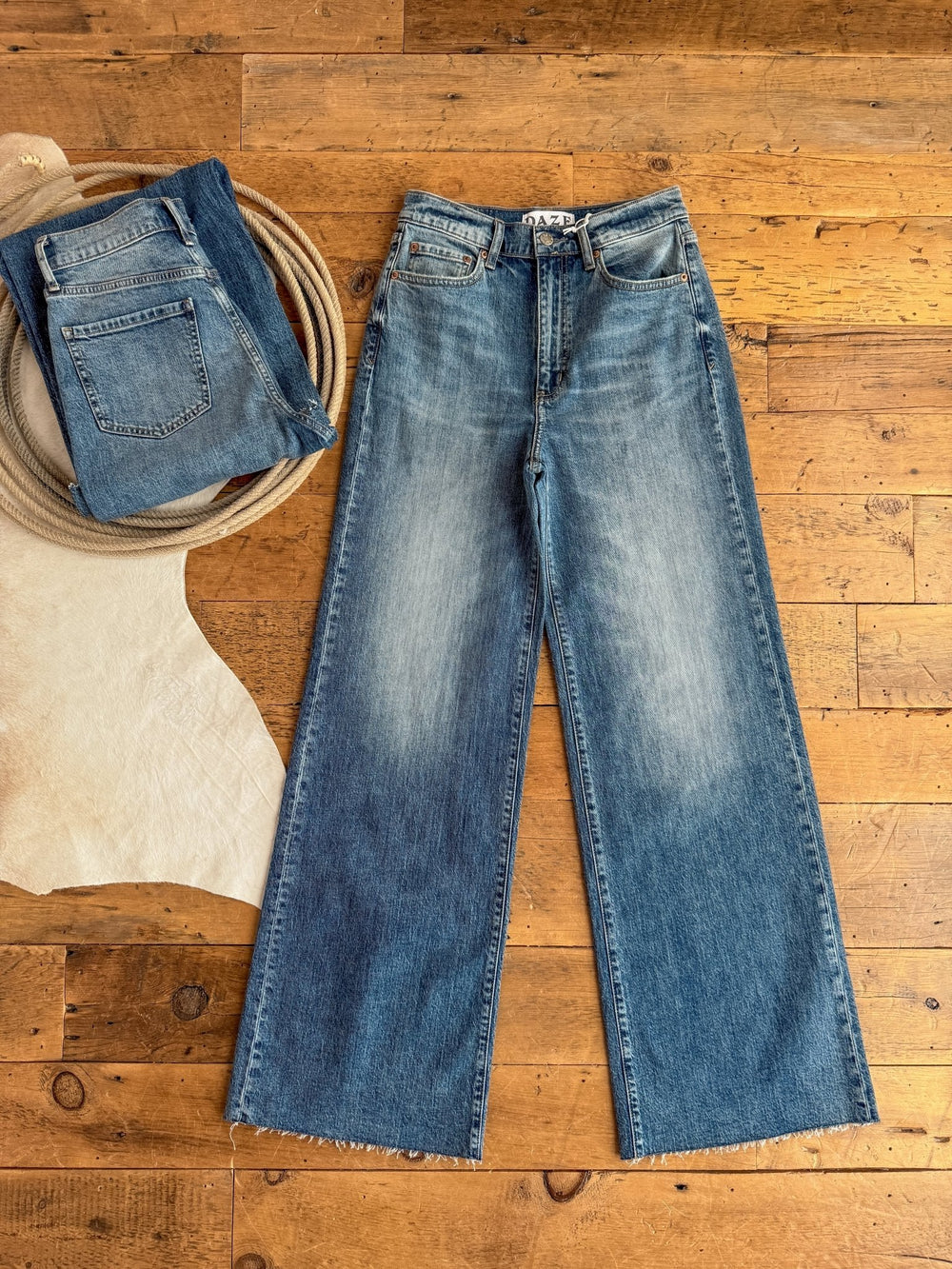 The Aiden High Rise Wide Leg Jean by Savannah Sevens western life{&}style is displayed flat on a wooden floor beside a folded pair of jeans, a coil of rope, and part of a light-colored animal hide visible on the left.