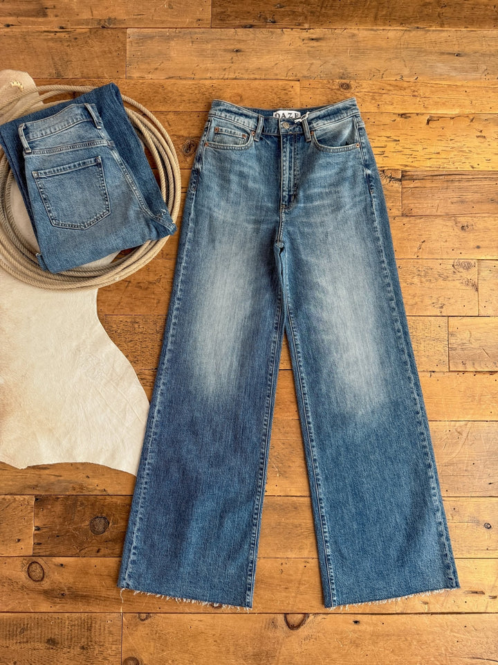 The Aiden High Rise Wide Leg Jean by Savannah Sevens western life{&}style is displayed flat on a wooden floor beside a folded pair of jeans, a coil of rope, and part of a light-colored animal hide visible on the left.